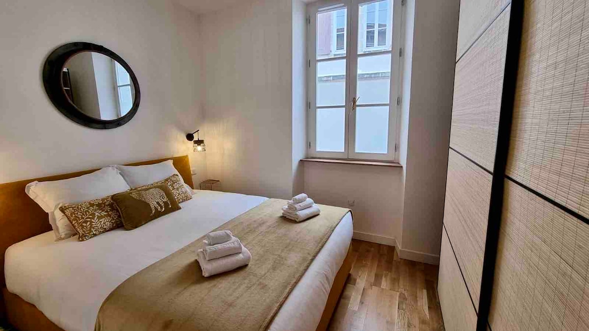 A cozy bedroom is illuminated by natural light from a window with a white frame. The spacious bed is dressed with neutral-colored linens and complemented by decorative pillows. A round mirror hangs above a bedside table, and a light fixture adds soft lighting. A wardrobe is visible against the wall.