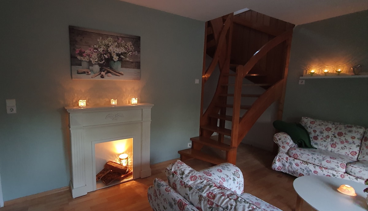 A cozy living area features floral-patterned sofas arranged around a simple white fireplace, where soft light flickers. A piece of wall art depicting flowers hangs above the mantel, while a wooden staircase leads to a second level, enhancing the space's inviting feel.