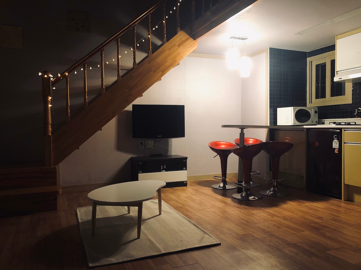 A cozy living area features a simple white coffee table resting on a soft rug. Below wooden stairs, a television stands, while a compact dining table with four bar stools is illuminated by pendant lights. The kitchen area is visible in the background.