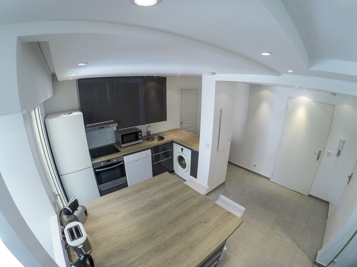 A modern kitchen is presented, featuring sleek cabinetry and stainless steel appliances. A spacious table is positioned at the center, surrounded by chairs. Natural light enters through a window, illuminating the area. An electric washer and essential kitchen equipment are clearly visible.