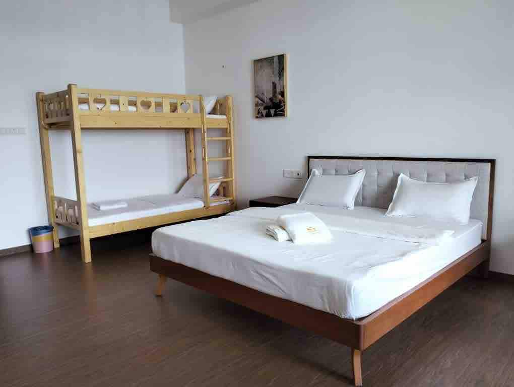 A bright and airy guest room features a spacious bed alongside a wooden bunk bed. The room is decorated with minimal furnishings, including a small artwork on the wall. Natural light fills the space through a large window, complementing the warm wood tones of the flooring.