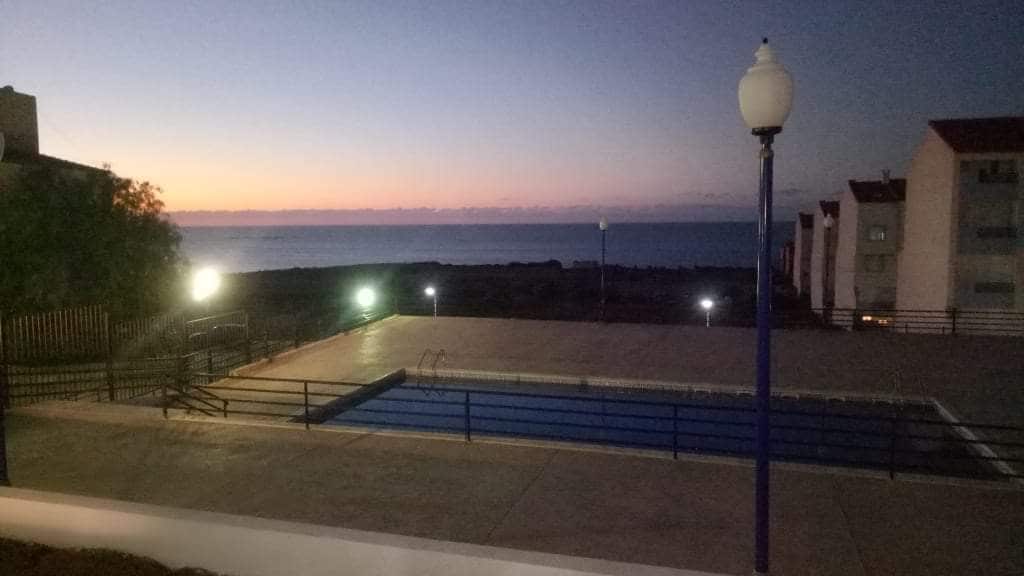 An evening view reveals a swimming pool surrounded by a concrete patio, with the calm sea in the background. The sky displays soft hues of dusk as lights illuminate the area, enhancing the tranquil ambiance.