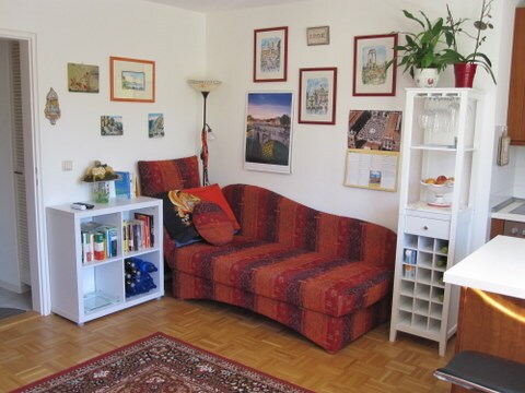 A cozy lounge area features a curved red patterned couch adorned with two cushions. A bookshelf and a white cabinet display books and decorative items. Walls are decorated with framed artwork and photographs. A floor lamp provides ambient lighting, enhancing the inviting space.