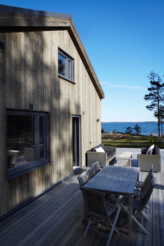 The exterior of the house features natural wood paneling and large windows, allowing for scenic views of the ocean. A spacious deck is equipped with a dining table and chairs, creating an inviting outdoor space for relaxation and enjoyment.