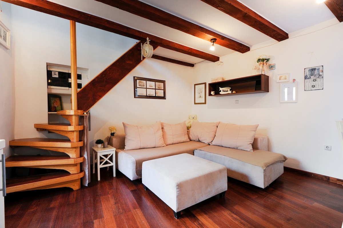 A spacious living area features a large L-shaped sofa with soft cushions and a light-colored ottoman. Wooden beams enhance the cozy atmosphere, while a spiral staircase adds an element of design. Shelves display decorative items, contributing to the welcoming environment.