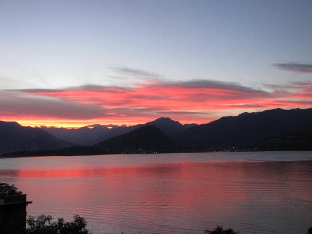 A serene sunset over Lago Maggiore is captured, displaying vibrant shades of red and orange against the horizon. The calm water reflects the colorful sky, while silhouettes of mountains create a tranquil backdrop, enhancing the peacefulness of the setting.
