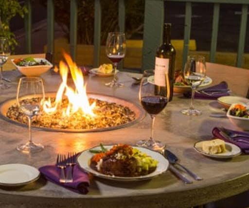 A circular dining table is set outdoors, featuring a central fire pit that flickers warmly. Plates of food are arranged alongside glasses of red wine and salad, with dark purple napkins folded neatly. The surrounding area is softly illuminated, creating an inviting ambiance.