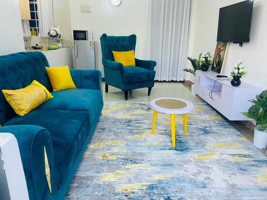 The living area features a vibrant blue couch adorned with yellow accent pillows. Two matching armchairs in a similar hue complement a light wooden coffee table, placed atop a decorative area rug with blue and yellow tones. A television is mounted on a sleek white media console.