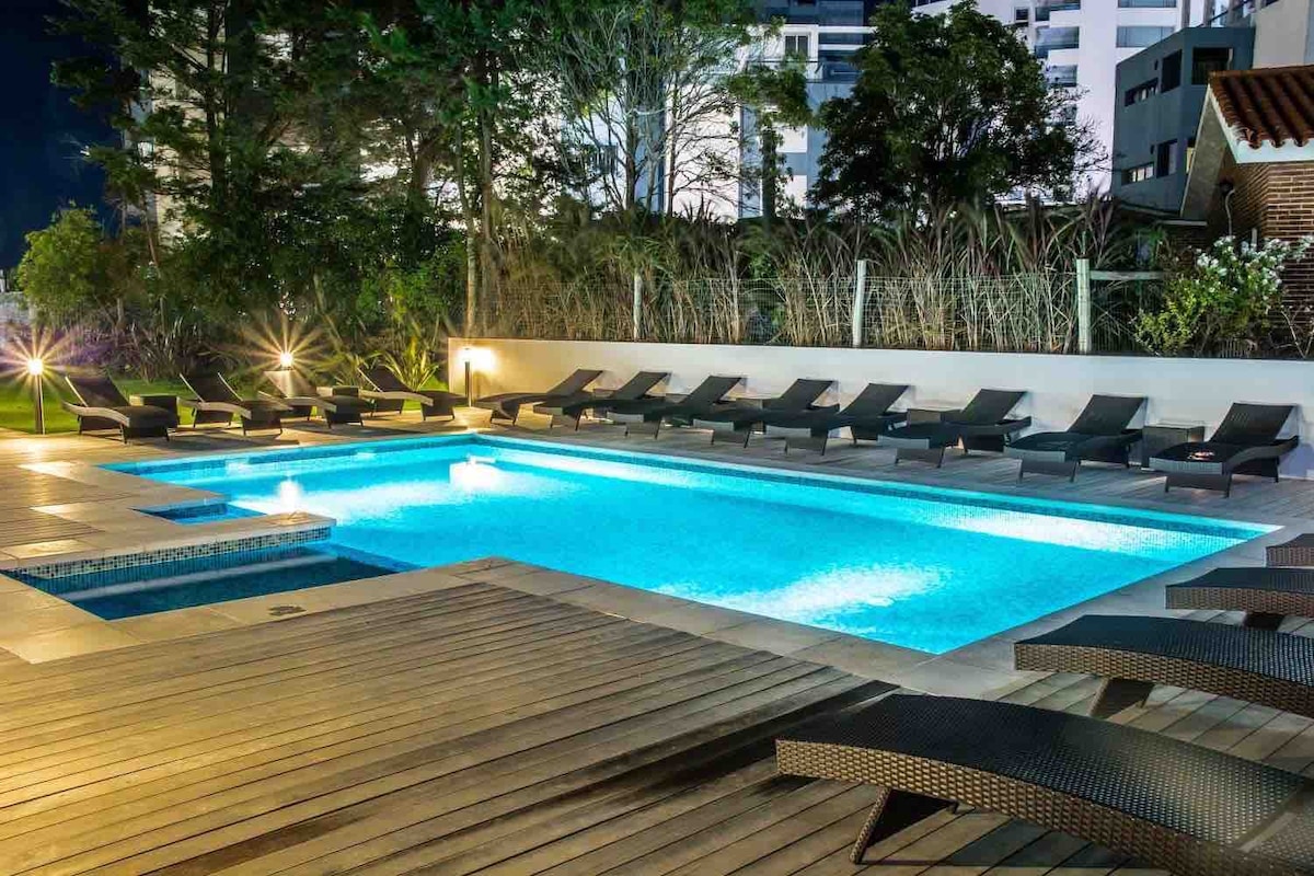 An outdoor heated swimming pool is surrounded by a wooden deck, featuring multiple lounge chairs arranged neatly. Soft lighting creates a calming atmosphere as greenery provides privacy around the pool area.