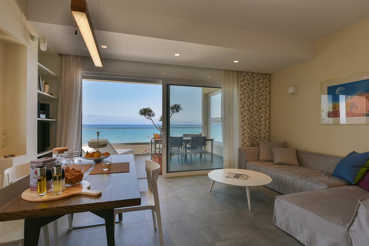 A bright living area features large glass doors offering unobstructed views of the sea. A dining table is set with refreshments, while a comfortable gray sofa accompanies a round coffee table. Natural light floods the space, highlighting the beach-inspired decor.