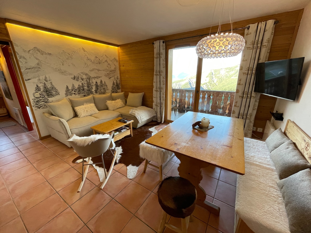 A cozy living area features a large sectional sofa accented with cushions, a wooden dining table, and a comfortable chair. Large windows offer scenic views of the mountains, while warm lighting enhances the wooden decor. A flat-screen TV is mounted on the wall.