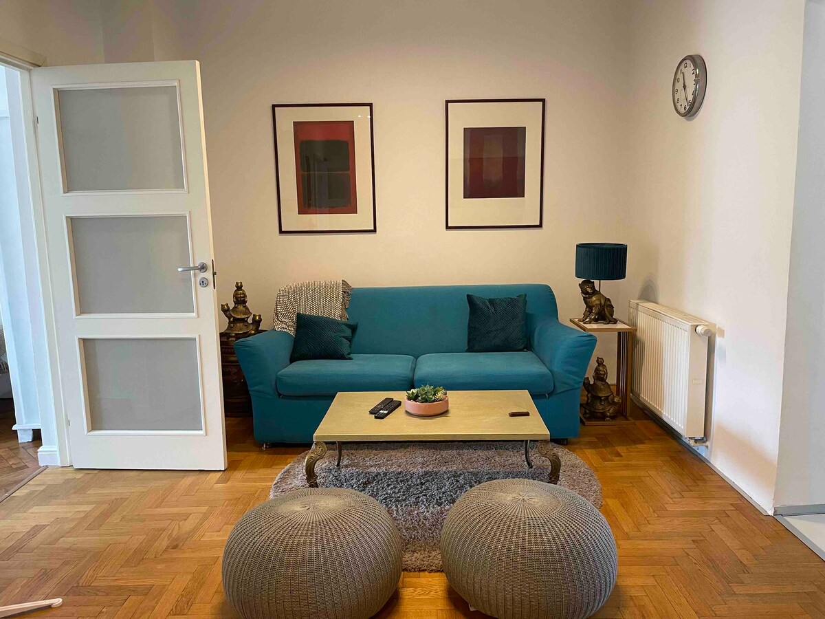 A living area is presented with a teal sofa paired with two round poufs. A wooden coffee table sits at the center, adorned with a small plant. Wall art pieces frame the space, complemented by a clock on the wall and a lamp beside the sofa.