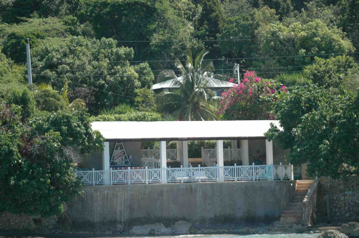 A covered outdoor entertainment space is framed by lush greenery and vibrant flowers. The structure features white railings and a sloped roof, providing shaded areas for relaxation. Steps lead down towards the water, with a serene atmosphere highlighted by the surrounding tropical landscape.
