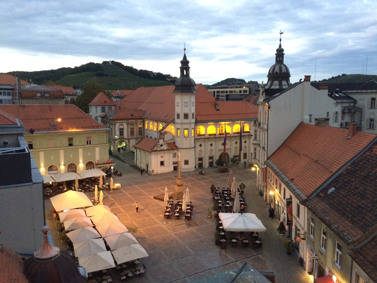 A view of a historic square in Maribor, featuring charming architecture and outdoor seating areas shaded by umbrellas. Soft evening light highlights the buildings, while the surrounding hills provide a serene backdrop. The space appears lively, with tables set for dining.
