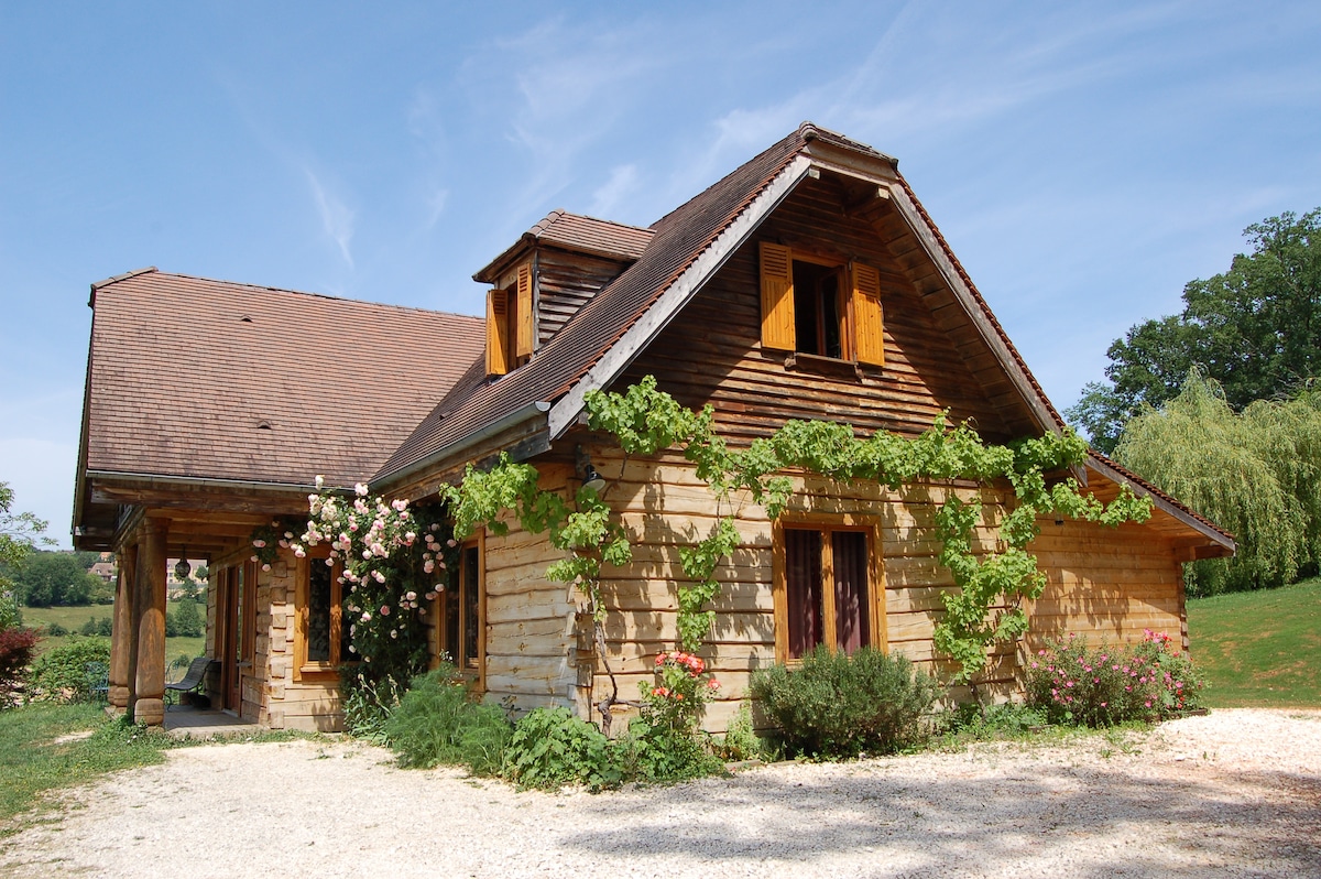 A wooden house is presented, adorned with climbing greenery and pink flowering plants. The structure features a charming sloped roof and multiple windows, allowing natural light to fill the space while a gravel path leads to the front entrance, surrounded by a lush lawn.