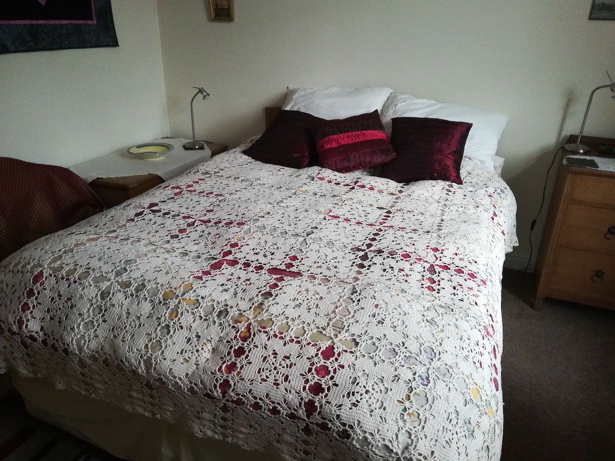 A spacious bed is covered with a decorative crochet blanket featuring intricate patterns in various colors. Two decorative pillows with a rich burgundy hue add a touch of elegance. A bedside lamp and a small table are positioned next to the bed, contributing to a cozy atmosphere.