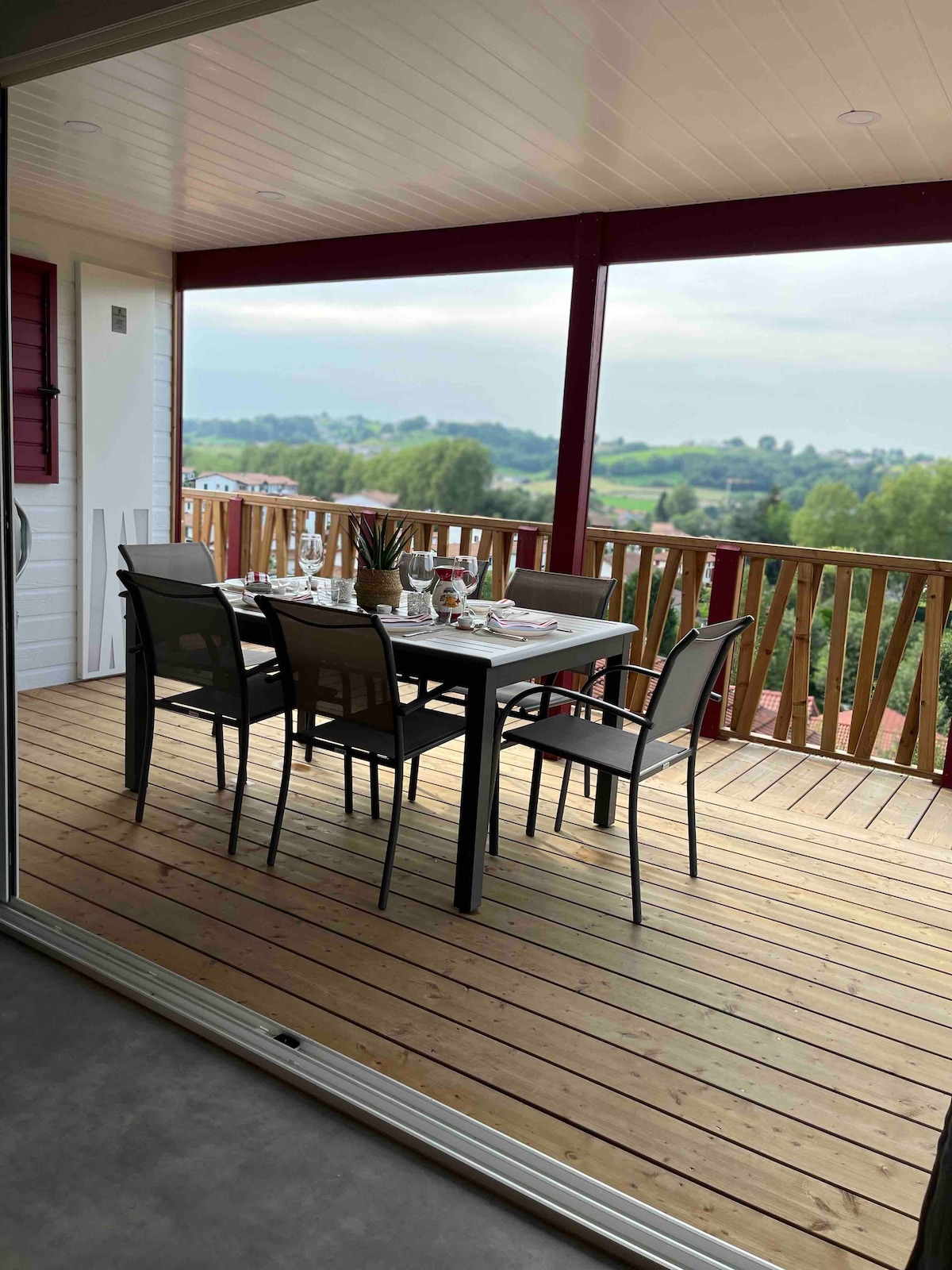 A dining area is situated on the terrace, featuring a table set for several guests. Black metal chairs surround the table, which is positioned on a wooden deck. The landscape beyond displays lush greenery and distant hills, complemented by a partly cloudy sky.