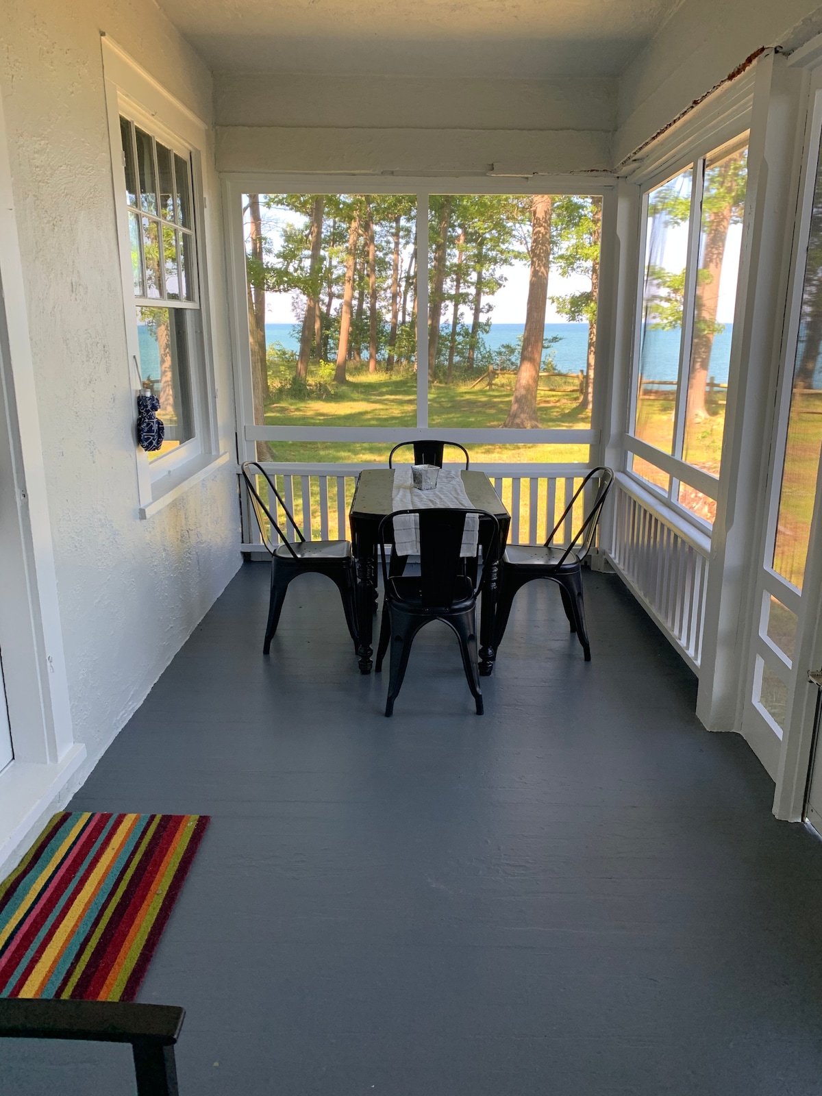 A screened-in porch features a dining table surrounded by four black chairs. Natural light floods the space, highlighting the wooden floors and white walls. Beyond the porch, views of tall trees and Lake Erie can be seen through the screens, creating a serene atmosphere.