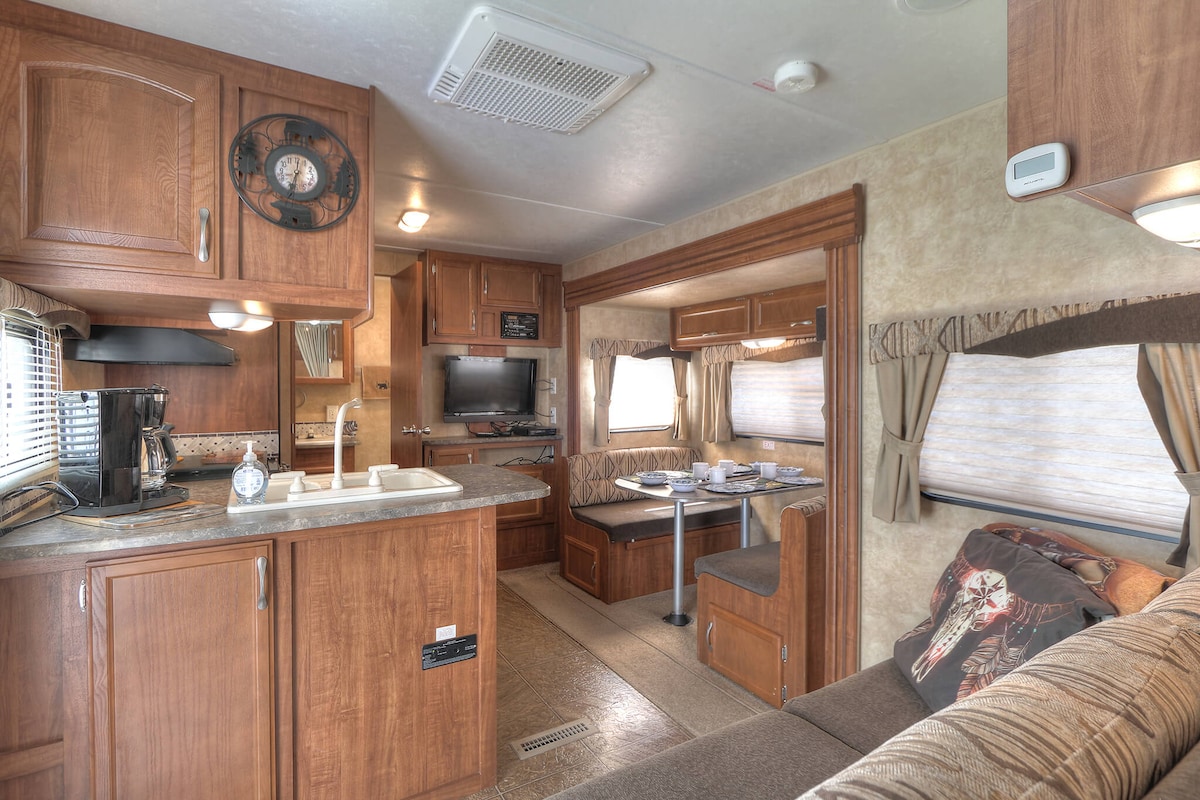 A well-equipped interior features wooden cabinetry and a cozy seating area. The kitchen displays essential appliances, including a coffee maker. A dining table is visible, flanked by comfortable seats. Natural light filters through the windows, enhancing the functional design of the space.