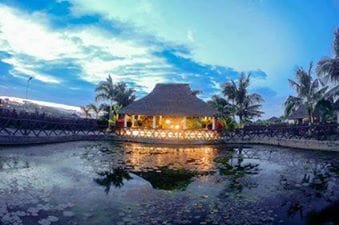 A traditional thatched-roof hut is surrounded by a calm pond, reflecting the evening sky. Palm trees frame the scene, and soft lighting from the hut creates a welcoming ambiance. The area is sheltered, offering a serene view of the water and surrounding greenery.