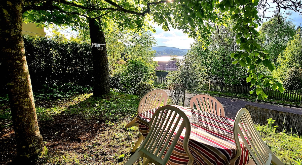 A shaded outdoor dining area is set beneath trees, featuring a table covered with a striped tablecloth and six white chairs. In the background, a serene lake view is visible, framed by lush greenery, creating an inviting environment for outdoor meals.