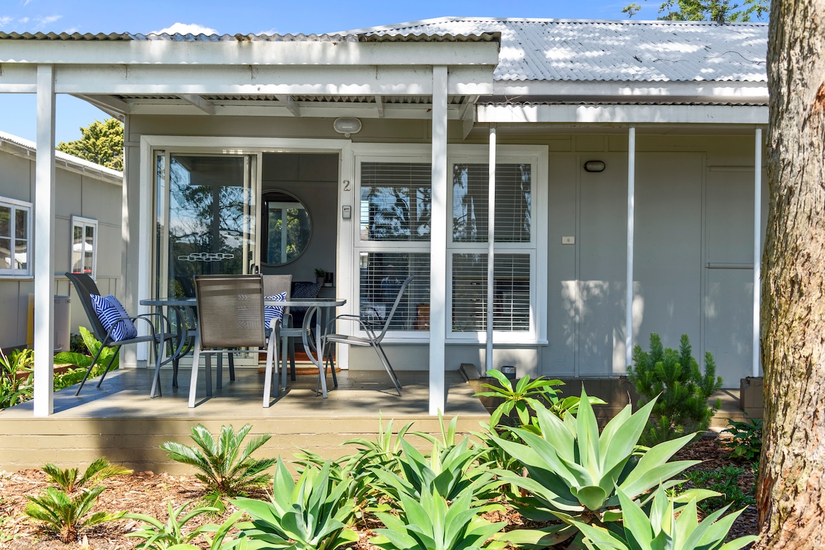 The exterior of Cottage No. 2 is depicted, featuring a covered private verandah furnished with a dining table and chairs. Large windows allow sunlight to brighten the space, while surrounding greenery adds a natural touch to the peaceful setting.