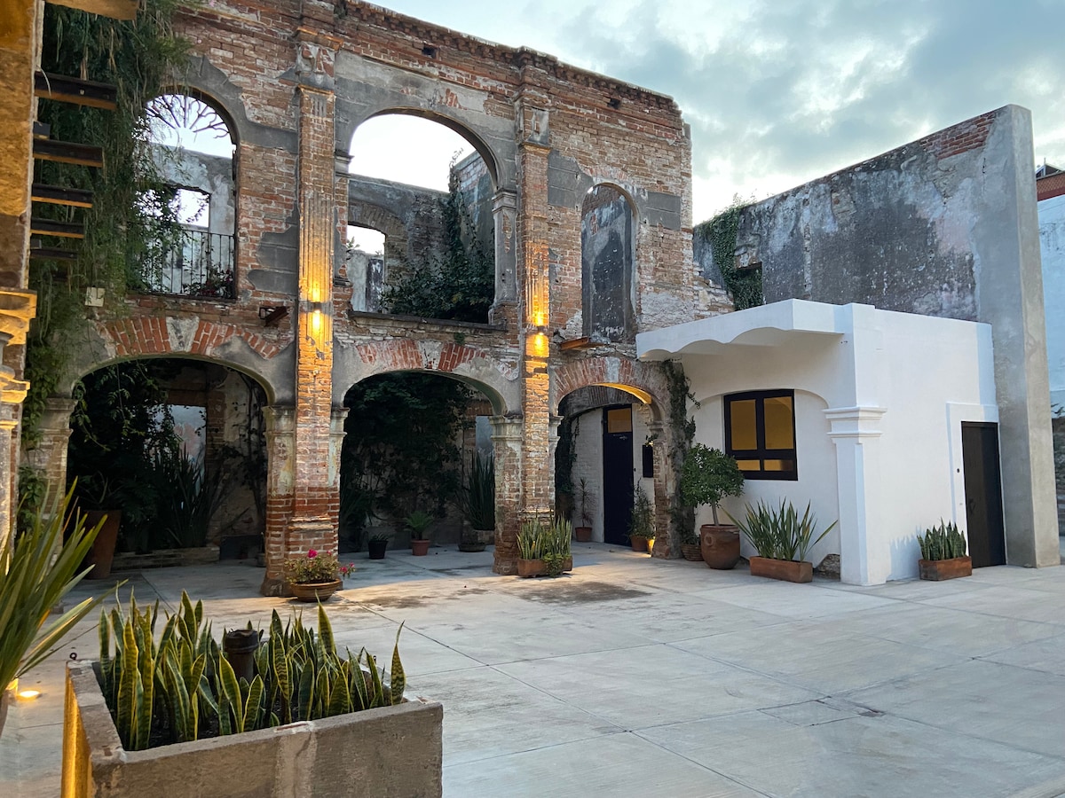 A historic building features exposed brick walls and arched windows, surrounded by plants in terracotta pots. Soft lighting enhances the architectural details, while an open courtyard area provides space for relaxation. The setting combines elements of antiquity and restoration.