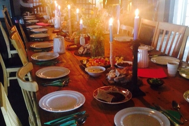A long wooden dining table is set with white plates and green cutlery, creating a welcoming dining environment. Candles flicker softly in the center, while bowls filled with an assortment of fruits and snacks are displayed, adding to the communal atmosphere.