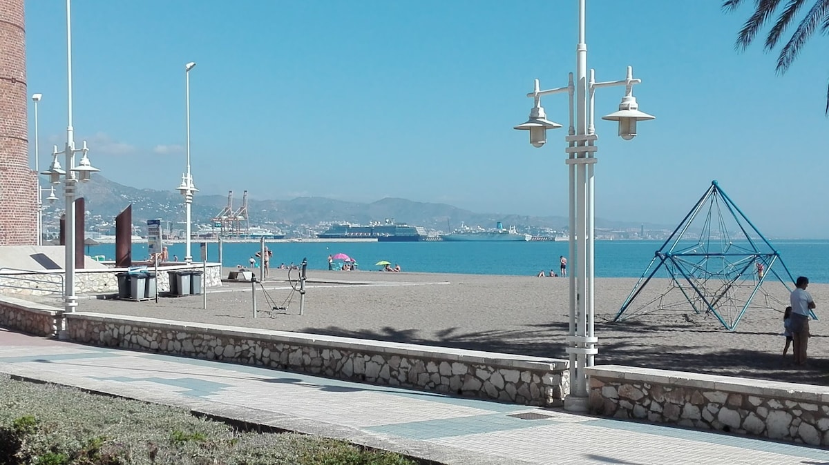 The image features a sandy beach extending towards the calm sea, with light waves lapping at the shore. In the foreground, a well-maintained promenade is observed, alongside inviting palm trees and a playful climbing structure for children. The distant horizon showcases ships on the water.
