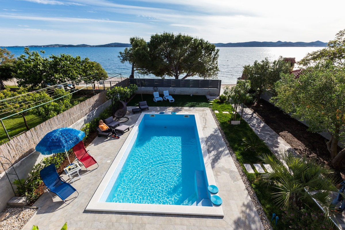 A peaceful outdoor area features a swimming pool surrounded by loungers and sun umbrellas. Lush greenery and trees create a natural boundary, while the beach appears in the background under a clear sky, offering a view of the calm sea.