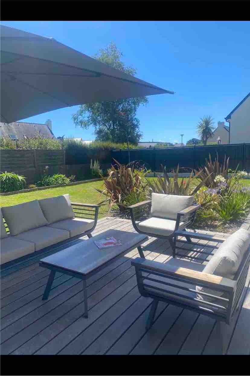 An outdoor seating area is presented on a wooden deck, featuring a table surrounded by comfortable chairs and a sofa. Lush greenery and garden plants create a serene backdrop, with a large umbrella providing shade from the sun.