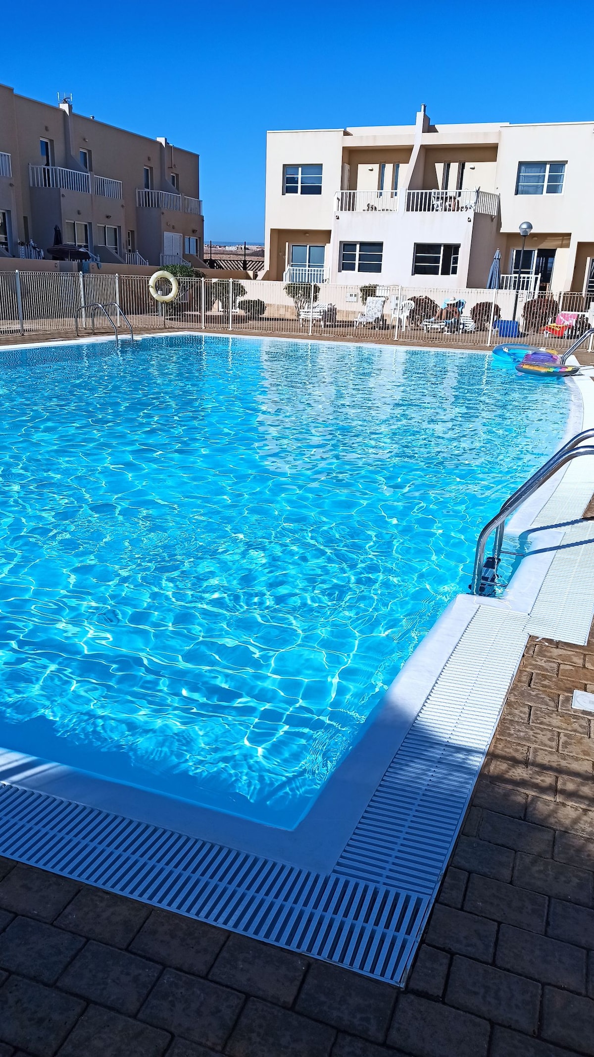 A large swimming pool is seen, featuring clear blue water that reflects the sunlight. Surrounding the pool are loungers and umbrellas, creating a relaxing environment. The area is paved with stone, and several nearby buildings are visible, contributing to a well-maintained setting.