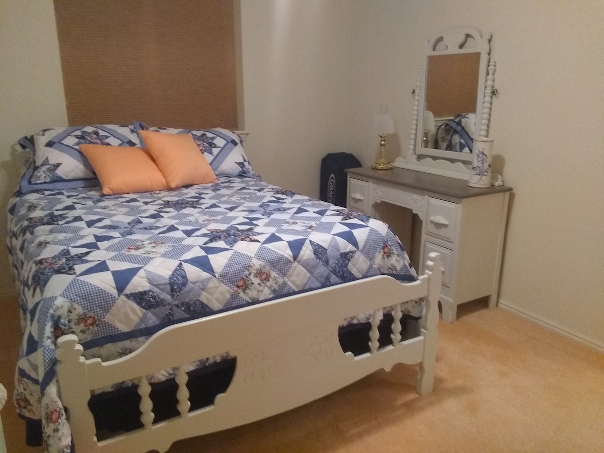A comfortable bedroom features a standard-sized bed dressed in a blue and white quilt with decorative pillows. A matching dresser with a mirror is positioned nearby, complementing the light-colored walls and soft carpet underfoot.
