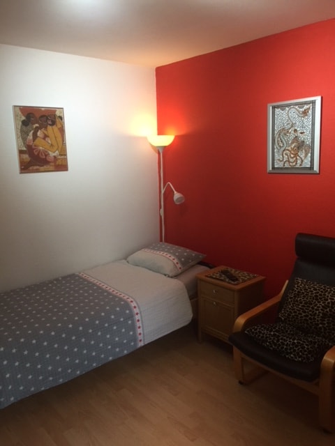 A compact bedroom features a single bed dressed in a light-colored duvet with subtle patterns. A warm light source illuminates the room. A wall-mounted lamp provides additional lighting, while a small nightstand holds a decorative item. A comfortable armchair complements the space.