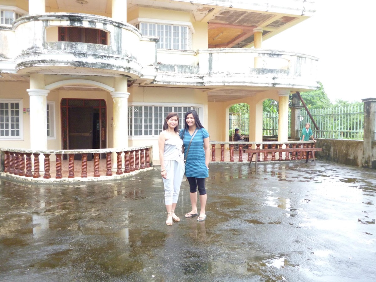 A spacious outdoor area features a large, two-story building with a covered porch. The ground is wet, indicating recent rain, and two individuals are positioned in front, dressed casually. The surrounding greenery is visible in the background, adding a natural element to the setting.