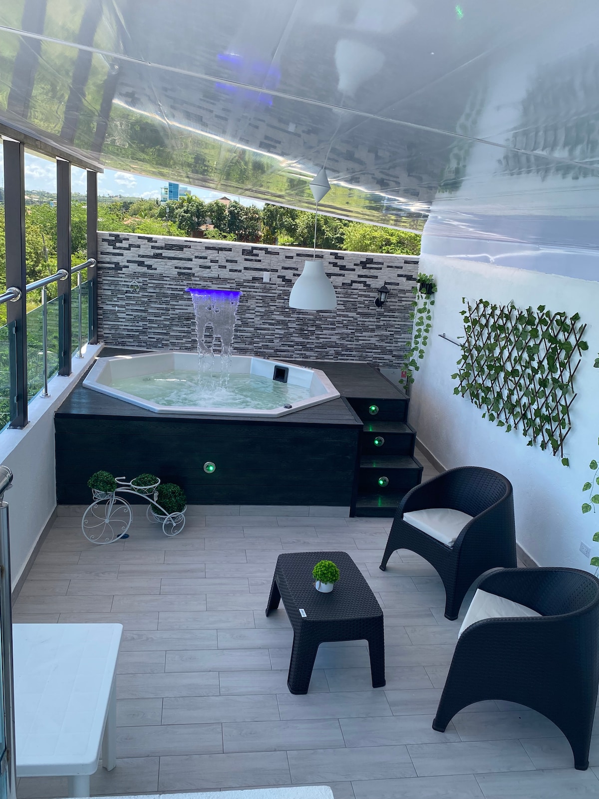 An octagonal Jacuzzi is situated in the corner, surrounded by stylish stone walls and greenery. Two black wicker chairs and a small table are positioned nearby, creating a relaxing seating area. Light streams in through large windows, enhancing the elegant decor.
