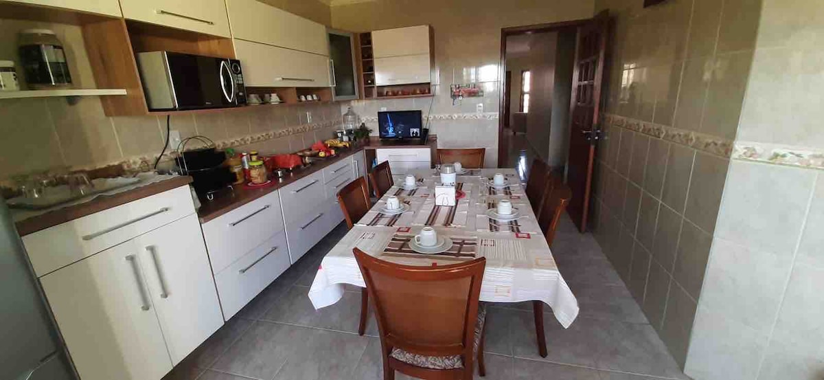A modern kitchen space features a long dining table set for six, with white tableware and napkins arranged neatly. The cabinets are light wood, complemented by white countertops. Appliances, including a microwave and coffee maker, are visible in the background, providing a functional cooking environment.