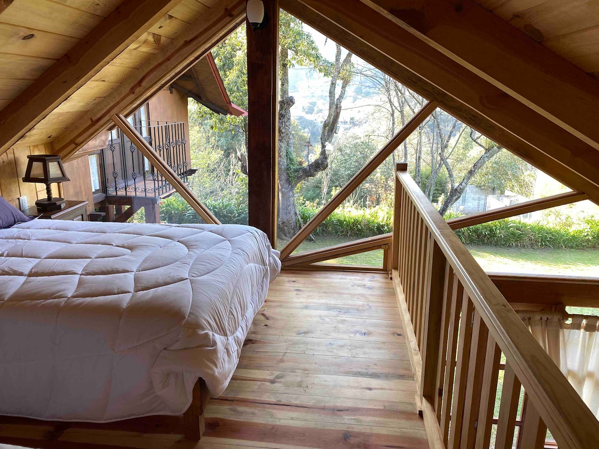 A well-lit sleeping area features a spacious bed, covered with a light comforter. The design incorporates wooden beams and large windows, allowing for a view of the garden and surrounding trees. A soft glow fills the space, creating a serene atmosphere.