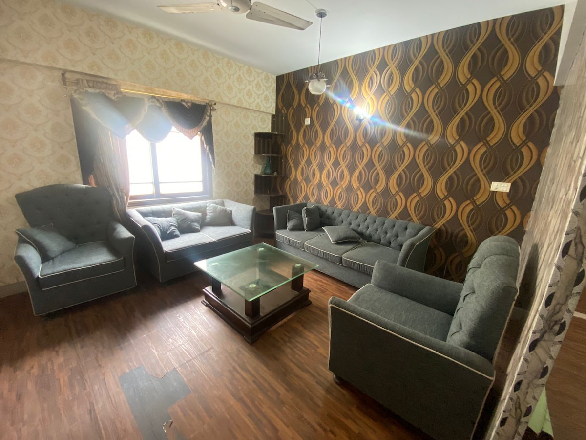 A comfortably arranged living area features a mix of upholstered seating, including a large sectional sofa and two armchairs. A glass coffee table sits in the center, complemented by patterned wallpaper and a large window that allows natural light to illuminate the space.