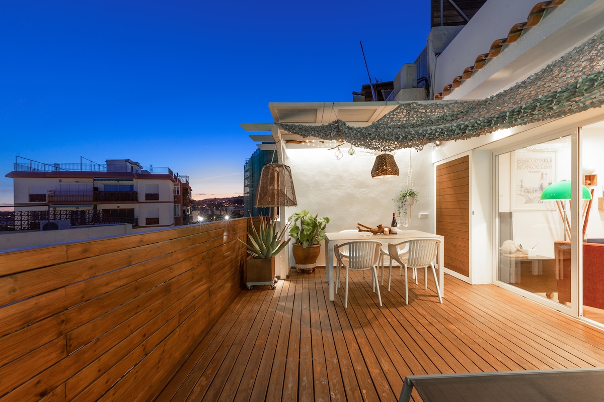 An outdoor terrace is featured with wooden decking, a dining table surrounded by chairs, and decorative pendant lights hanging overhead. Potted plants are visible, and a warm evening sky in shades of blue serves as a backdrop, enhancing the serene outdoor setting.