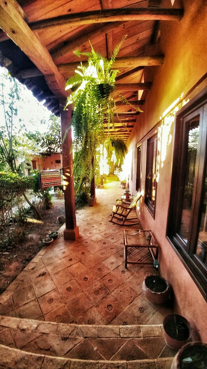 A shaded outdoor corridor with a tiled floor features wooden beams overhead. Potted plants and ferns add greenery along the walking path. Two wooden rocking chairs provide a quiet seating area. Natural light filters through the space, creating a warm ambiance.