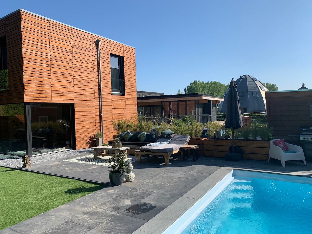 A modern wooden villa is embraced by lush greenery, showcasing a private swimming pool. Outdoor lounge beds are positioned near the water, complemented by a shaded area with a large umbrella. The clean lines of the architecture enhance the serene outdoor space.