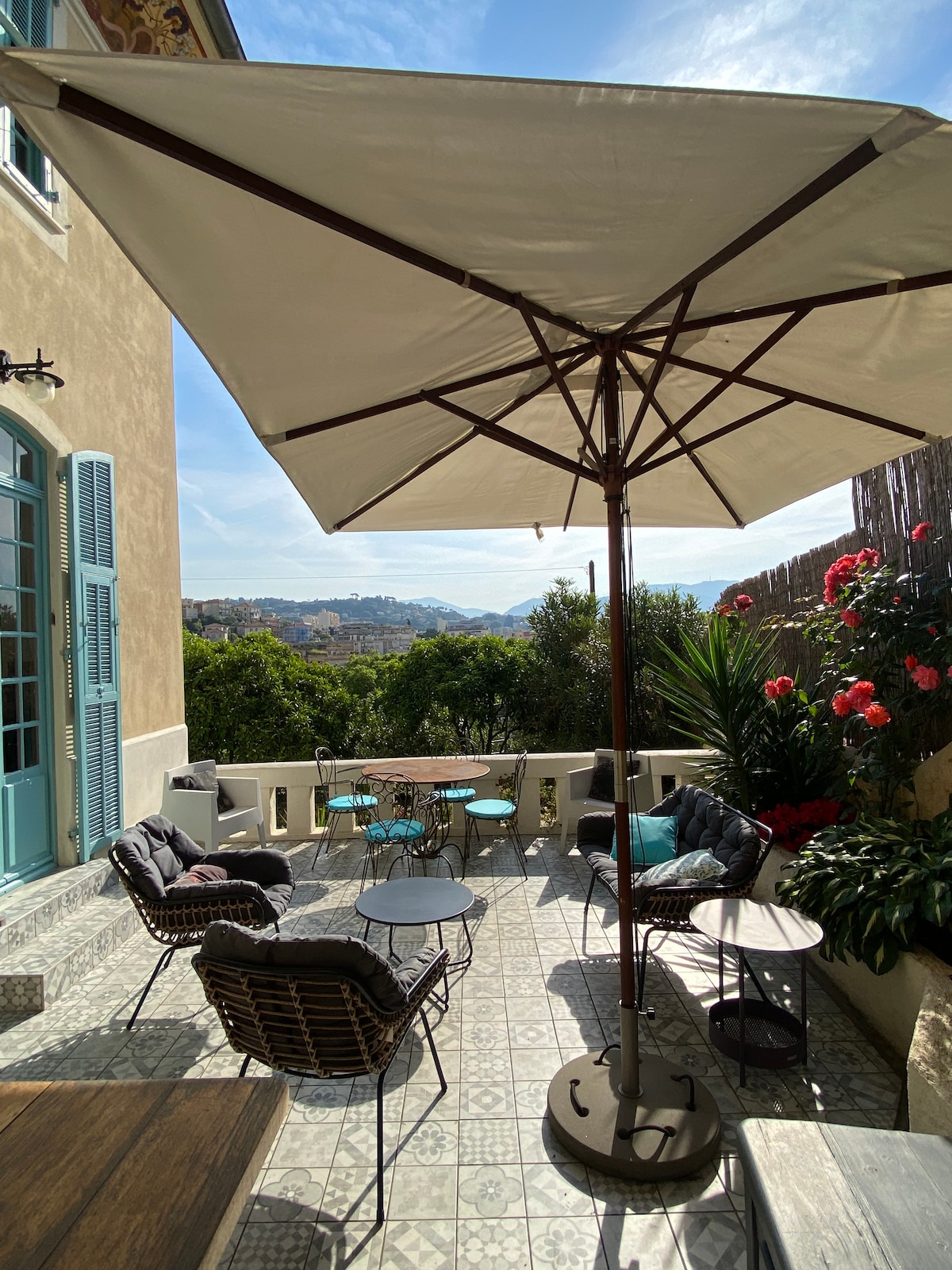 An outdoor terrace features a round table surrounded by soft seating, sheltered by a large umbrella. Lush greenery and flowering plants enhance the space, while scenic views of the hills are visible in the background. A delicate tile pattern covers the terrace floor.