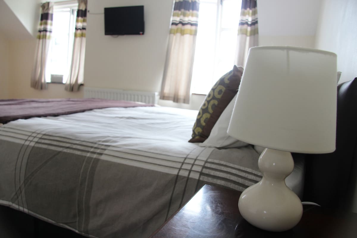 A spacious room is depicted, featuring a large bed with layered bedding. A modern lamp is placed on a dark wooden bedside table. A flat-screen TV is mounted on the wall, and light filters in through two curtained windows.