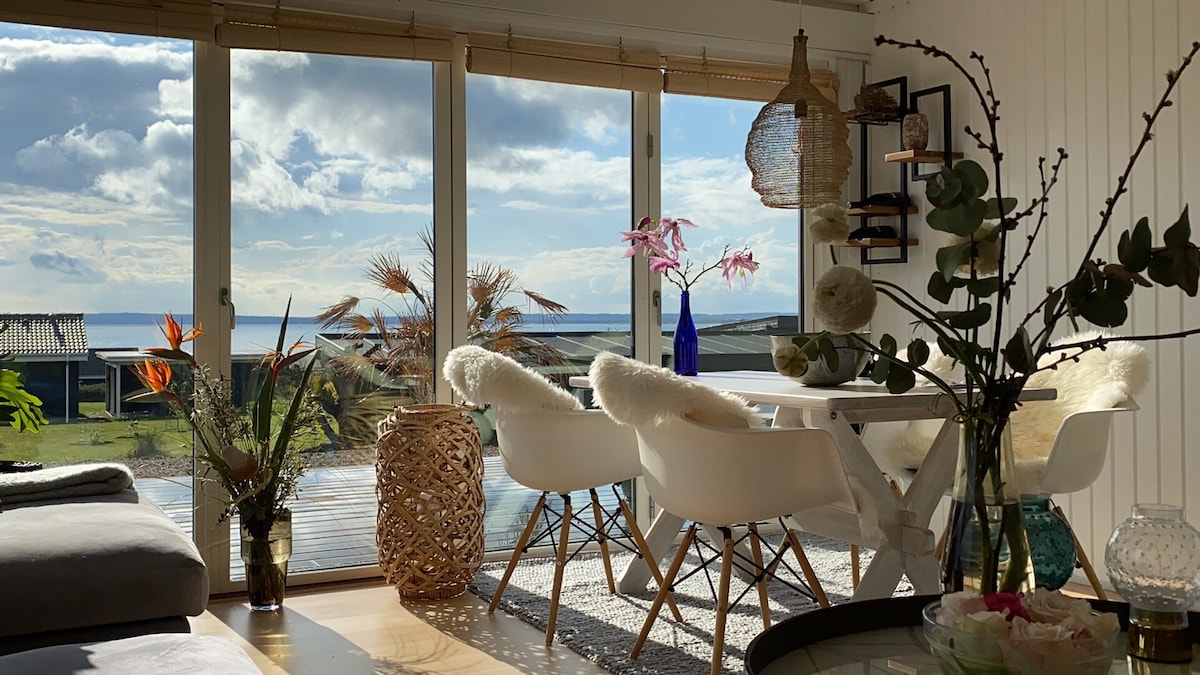 A dining area is shown, featuring a white table surrounded by four armchairs. Natural light pours through expansive sliding glass doors that offer views of the sea and sunny skies. Decorative plants and a vase with flowers add touches of greenery and color to the space.