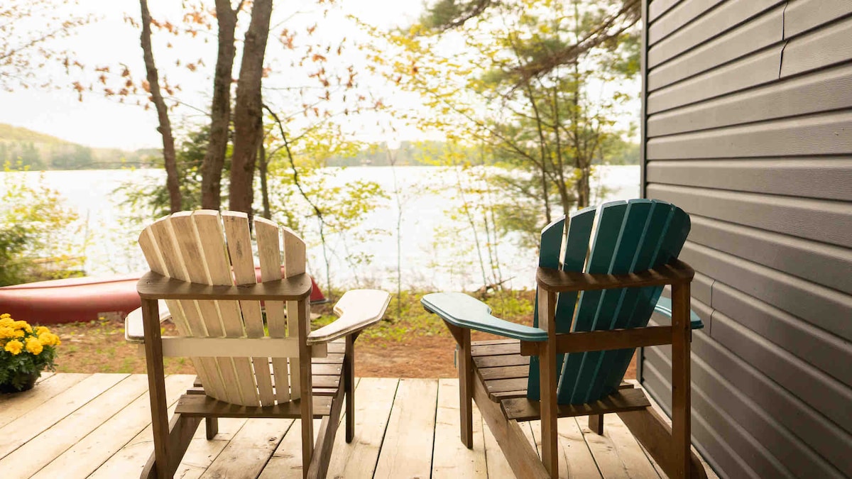 Two Adirondack chairs are positioned on a wooden deck, facing a serene lakeside view. Soft greenery surrounds the area, complementing the tranquil atmosphere. A canoe is visible by the shore, enhancing the connection to nature.