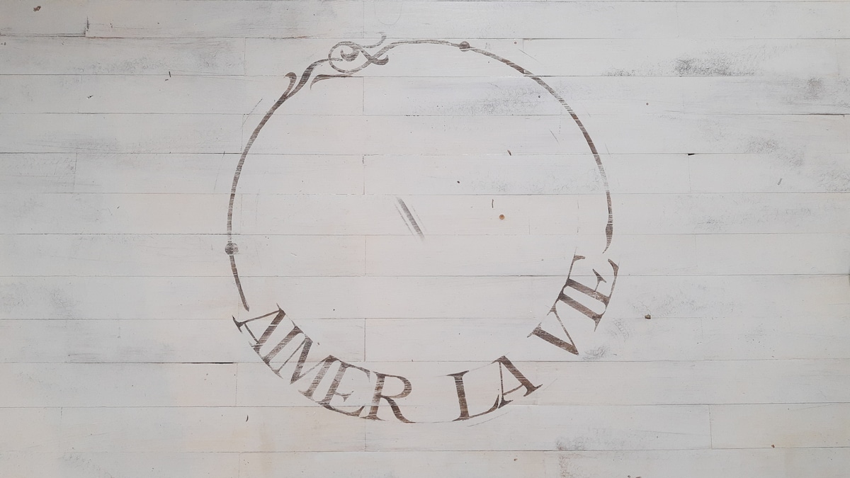 A circular design featuring the phrase 'AIMER LA VIE' is displayed on a whitewashed wooden floor, enhancing the room's character. The light color of the wood creates a bright and open feel in the space.