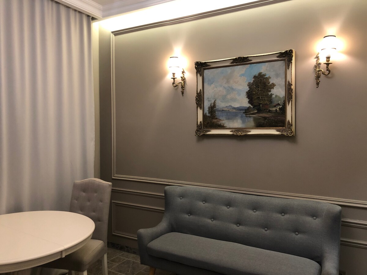A light-colored wall is adorned with an elegant painting, depicting a serene landscape. Soft lighting is provided by two sconces on either side. A round dining table is paired with two light fabric chairs, and a subtle gray sofa adds a touch of comfort.