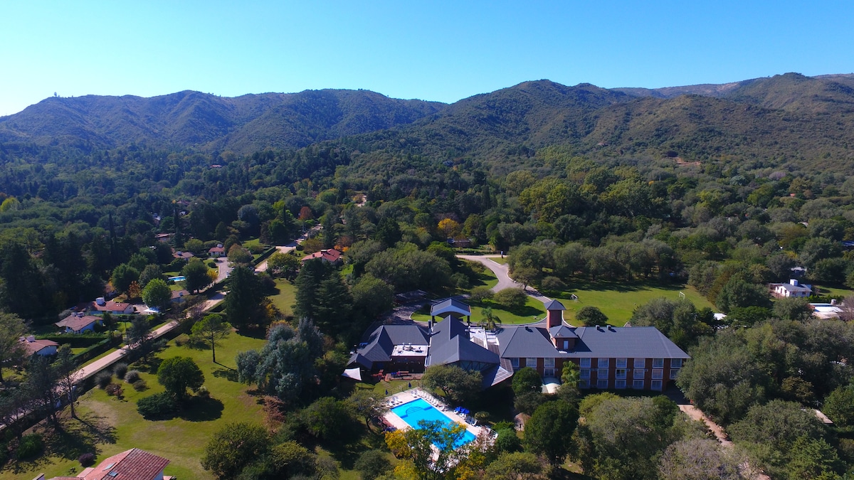 An aerial view captures the hotel surrounded by lush greenery and rolling hills. A clear blue pool is positioned in the garden area, reflecting the vibrant landscape. The building's architecture complements the natural surroundings, creating an inviting atmosphere.