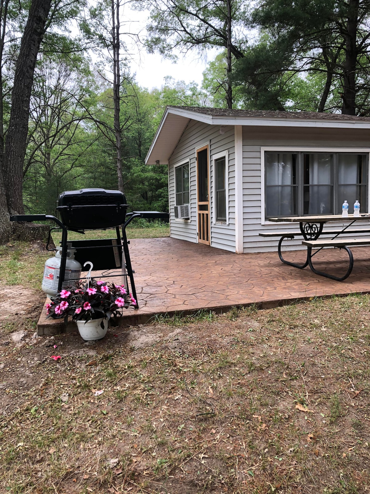 A charming patio area features a black gas grill next to a dining table with a circular design. Potted pink flowers add a touch of color, while the cottage is visible, surrounded by tall trees that provide a natural backdrop.